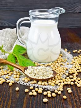 Soy flour in a spoon and milk in glass jug, soybeans and a leaf on burlap on the background of wooden board
