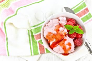 Ice cream crimson with raspberry berries, syrup and mint in white bowl, a spoon on towel against light wooden board on top