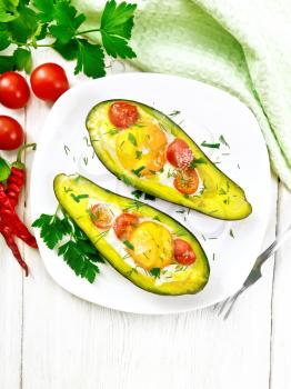 Scrambled eggs with cherry tomatoes in two halves of avocado in a plate, napkin and fork on a background of white wooden board from above