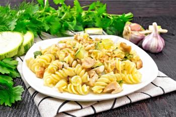 Fusilli pasta with chicken breast, zucchini, cream and pine nuts in a plate on towel, garlic, fork and parsley on a wooden board background