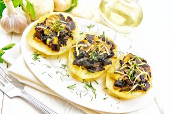 Potatoes stuffed with mushrooms, fried onions and cheese in a plate on towel, vegetable oil in decanter, parsley, garlic and a fork on the background of light wooden board