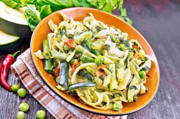 Tagliatelle pasta with zucchini, green peas, asparagus beans, hot peppers and spinach in a plate on towel, garlic, fork and basil on wooden board background