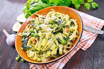 Tagliatelle pasta with zucchini, green peas, asparagus beans, hot peppers and spinach in a plate on napkin, garlic, fork and basil against dark wooden board