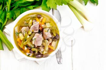 Eintopf soup of pork, celery, beans, carrots and potatoes with leek in a white bowl on a napkin on wooden board background from above