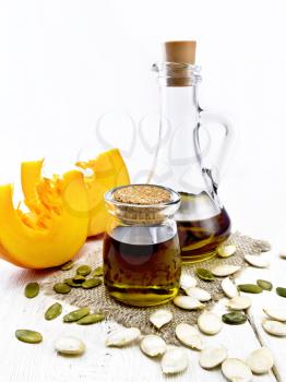 Pumpkin oil in a jar and decanter on burlap, seeds on the table, slices of vegetable on background of light wooden board
