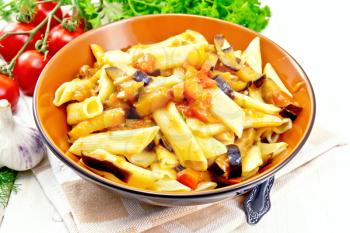 Penne pasta with eggplant and tomatoes in a bowl on kitchen towel, fork, garlic and parsley on light wooden board background