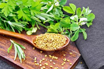 Fenugreek seeds in a spoon on a brown plate with spicy herbs, dark napkin on black wooden board background