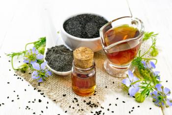 Nigella sativa oil in vial and gravy boat, seeds in a spoon and black cumin flour in a bowl on burlap, kalingi twigs with blue flowers and green leaves on wooden board background