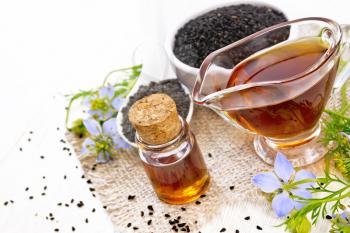 Nigella sativa oil in vial and gravy boat, seeds in a spoon and black cumin flour in a bowl on burlap, kalingi twigs with blue flowers and leaves on white wooden board background