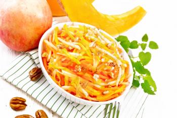 Pumpkin, carrot and apple salad with pecans seasoned with vegetable oil in a bowl on a napkin, mint on light wooden board background