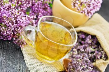 Oregano herbal tea in a glass cup on burlap napkin, fresh flowers in mortar and on the table, dry marjoram flowers in a bag and spoon on dark wooden board background