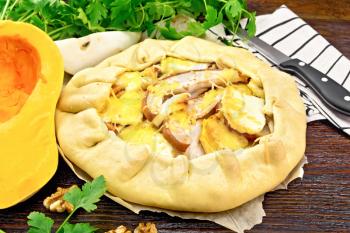 Pie of pumpkin, pear, soft cheese and walnuts on paper, napkin and knife, parsley on a dark wooden board