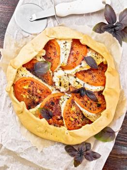 Pie with tomatoes, curd cheese and purple basil on parchment on a dark wooden board background from above