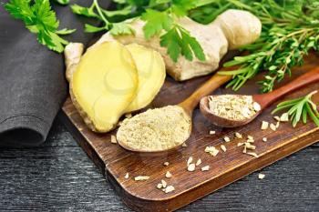 Ground ginger and flakes in two wooden spoons, ginger root, spicy herbs, napkin on a black wooden board background