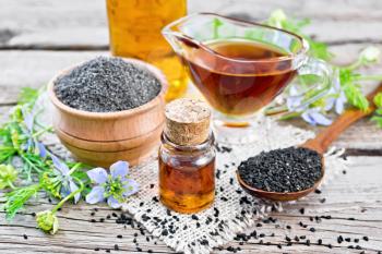 Nigella sativa oil in vial, gravy boat and bottle, seeds in a spoon and black cumin flour in a bowl on burlap, kalingi twigs with blue flowers and green leaves on wooden board background