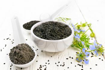 Black cumin seeds in a bowl, flour and seeds in spoons, kalingi sprigs with blue flowers and green leaves on a wooden board background