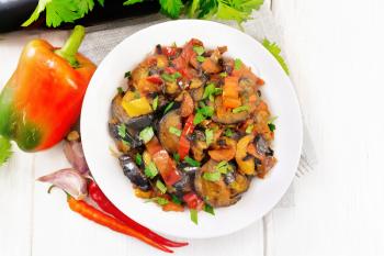 Vegetable ragout with eggplant, tomatoes, sweet and hot peppers, onions, carrots, fried with herbs and spices in plate on a towel, garlic, parsley on the background of light wooden board from above