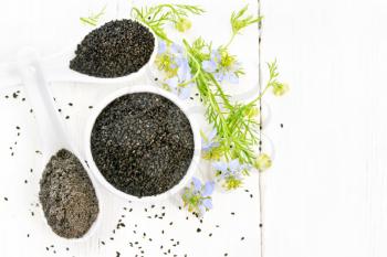 Black cumin seeds in a bowl, flour and seeds in spoons, kalingi sprigs with blue flowers and green leaves on a wooden board background from above