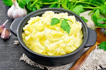 Mashed potatoes in a black saucepan and a spoon on a burlap napkin, garlic, parsley on wooden board background