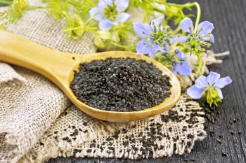 Black cumin seeds in a spoon on sacking, kalingi twigs with blue flowers and green leaves on a dark wooden board background