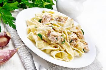 Tagliatelle pasta with salmon, cream, garlic and herbs in a plate on a napkin, fork, parsley and basil on white wooden board background