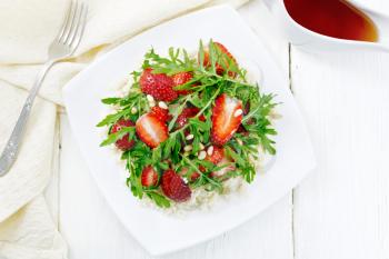 Strawberry, couscous, cedar nuts and arugula salad dressed with balsamic vinegar and olive oil in a plate, napkin and fork on wooden board background from above