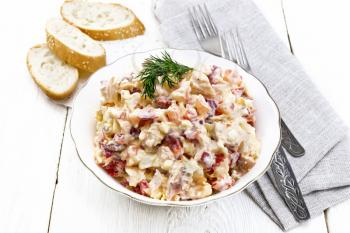 Salad with chicken, sweet pepper, tomato, egg and cheese seasoned with mayonnaise and garlic in a dish, gray napkin, bread and forks on a wooden board background