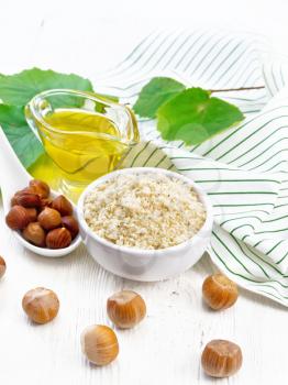 Hazelnut flour in a bowl, oil in a glass gravy boat, nuts, napkin and filbert branch with green leaves on wooden board background