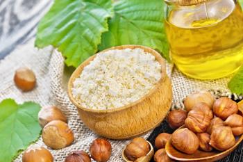 Hazelnut flour in a bowl, nuts in a spoon, oil in glass jar and filbert branch with green leaves on burlap on wooden board background
