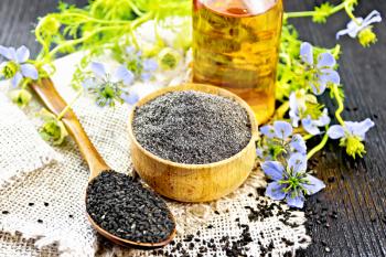 Flour of black caraway in a bowl, seeds in a spoon on burlap, oil in bottle and twigs Nigella sativa with blue flowers and leaves on black wooden board background