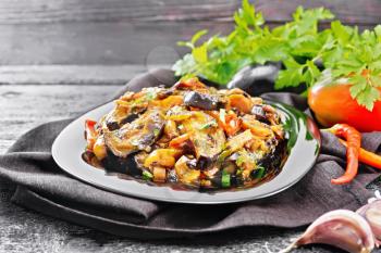 Vegetable ragout with eggplant, tomatoes, sweet and hot peppers, onions, carrots, fried with herbs and spices in a plate on kitchen towel, garlic, parsley on wooden board background