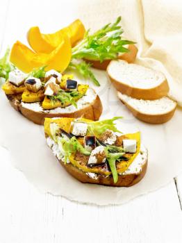 Bruschetta with baked pumpkin, salted feta cheese, ricotta, arugula, spices and balsamic sauce on parchment, napkin on white wooden board background