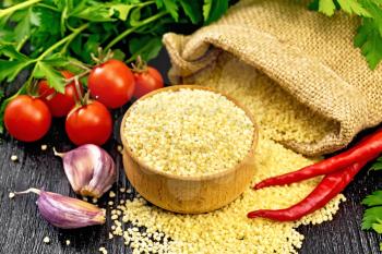 Raw couscous in a bowl and a sack of burlap, tomatoes, hot peppers, herbs and garlic on wooden board background