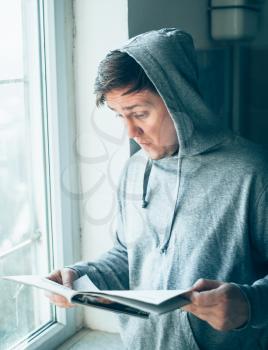 A man in hood looks at a magazine near the window. Press hands.