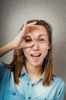 Young woman watching through fingers okay glasses over a grey background