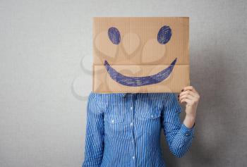 Young woman holding a cardboard with a smiley face on it in front of her head.