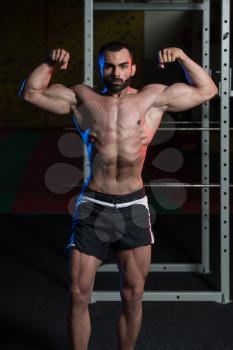 Young Man Standing Strong In The Gym And Flexing Front Double Biceps Pose - Muscular Athletic Bodybuilder Fitness Model Posing Exercises