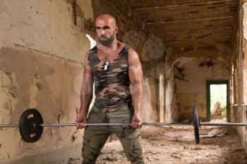 Handsome Muscular Fitness Man In Army Pants Doing Heavy Weight Exercise For Biceps With Barbell Inside Shelter