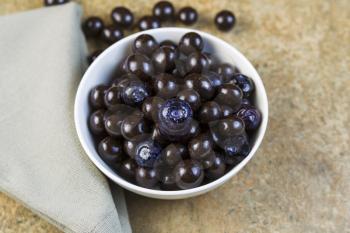 Horizontal photo, focus on large blueberry in center, of blueberries and chocolate balls in small white bowl with light green cloth napkin on natural slate as background
