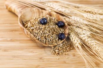 Closeup of horizontal photo of wooden spoon filled with Whole Grain Cereal with blueberries on top, wheat stalks on side with natural bamboo wood in background