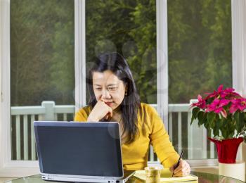 Mature Asian woman working at home with notebook computer on glass table with large windows in background
