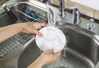 Horizontal photo of female hands rinsing off a small white dinner plate with kitchen sink and running faucet in background