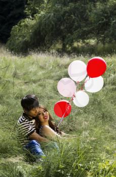 Vertical photo of young adult couple sitting in the middle of green tall grass field  with balloons and trees in background 