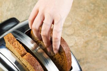 Horizontal photo of female hand putting fresh whole wheat bread into toaster with stone counter top in background 