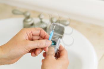 Horizontal photo of female hands holding dental tooth pick in bathroom with dental floss container, sink and counter top in background 