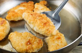 Horizontal photo of golden breaded coated fish being fried in stainless steel frying pan with focus on single piece with spatula underneath