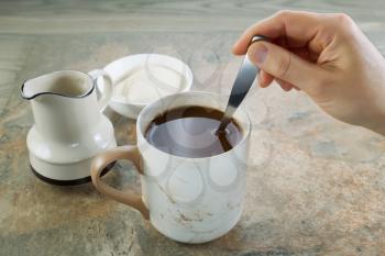 Closeup horizontal photo of female hand stirring cream and sugar with spoon in a cup of black coffee with stone and aged wood underneath
