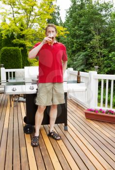 Vertical photo of mature man drinking a beer out of a glass with barbecue cooker and seasonal trees in background 