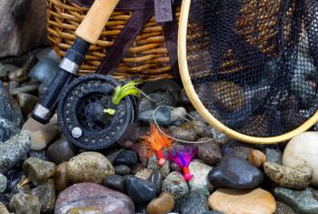 Closeup of fishing fly reel, landing net, creel and assorted flies on wet river bed stones
