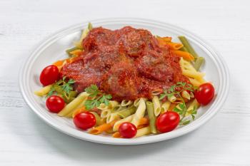 Freshly cooked penne pasta, homemade meatballs, cherry tomatoes and parsley on white plate with white table as background. 
